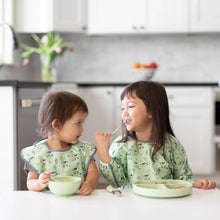 Load image into Gallery viewer, Two kids in matching bibs eat a meal at a white table in a kitchen; one uses Bumkins Spoon + Fork: Sage.
