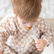 Load image into Gallery viewer, A boy in a tan checkered bib uses Bumkins Spoon + Fork: Sand to eat a meal in a high chair.
