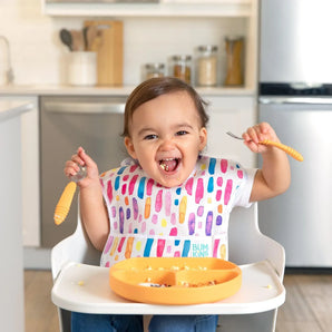 A happy toddler with a colorful bib sits in a high chair, holding Bumkins Spoon + Fork: Tangerine over a divided plate of food.