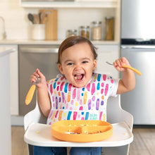 Load image into Gallery viewer, A happy toddler with a colorful bib sits in a high chair, holding Bumkins Spoon + Fork: Tangerine over a divided plate of food.

