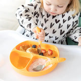 A toddler eats pancakes, fruit, and yogurt from an orange plate using Bumkins Spoon + Fork: Tangerine, wearing a heart-patterned bib.