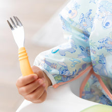 Load image into Gallery viewer, Close-up of a child in a pastel bib holding a Bumkins Spoon + Fork: Tangerine.
