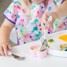 Load image into Gallery viewer, A child in a floral bib enjoys colorful food from a plate with Bumkins Spoon + Fork: Vanilla Sprinkle.

