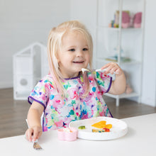 Load image into Gallery viewer, A smiling blonde girl in a floral bib uses Bumkins Spoon + Fork: Vanilla Sprinkle to eat from a white plate at a white table.
