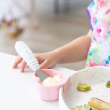 Load image into Gallery viewer, Close-up of a child using Bumkins Spoon + Fork: Vanilla Sprinkle to enjoy yogurt from a pink heart-shaped cup at a white table.
