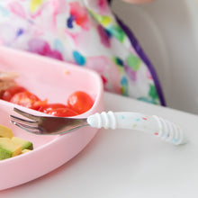 Load image into Gallery viewer, Close-up of a Bumkins Spoon + Fork: Vanilla Sprinkle resting on a pink sectioned plate with tomatoes and avocado on a white table.
