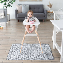Load image into Gallery viewer, A baby in a high chair sits on a Bumkins Splat Mat: Hearts, eating from a bowl. A gray sofa and decor adorn the modern living room behind.
