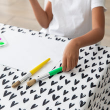 Load image into Gallery viewer, A child in a white shirt sits at a table covered with a Bumkins Splat Mat: Hearts, holding green and yellow brushes next to paper.
