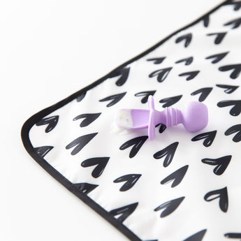 Close-up of a purple baby spoon with food sitting on a Bumkins Splat Mat: Hearts, made from easy-wipe fabric, on a white background.