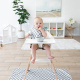 A baby in a high chair with a checkered bib enjoys a meal over the Bumkins Splat Mat: Charcoal Check, next to a plant and playhouse.