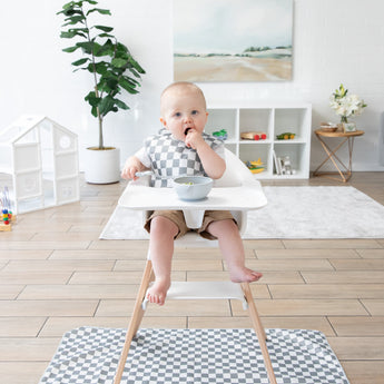 A baby in a high chair with a checkered bib enjoys a meal over the Bumkins Splat Mat: Charcoal Check, next to a plant and playhouse.