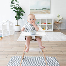 Load image into Gallery viewer, A baby in a high chair with a checkered bib enjoys a meal over the Bumkins Splat Mat: Charcoal Check, next to a plant and playhouse.
