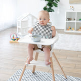A baby wearing a bib in a bright room with toys sits in a high chair with a Bumkins Splat Mat: Charcoal Check underneath, holding a spoon.