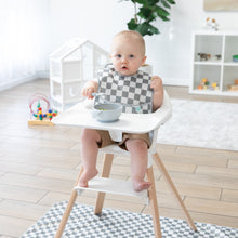 Load image into Gallery viewer, A baby wearing a bib in a bright room with toys sits in a high chair with a Bumkins Splat Mat: Charcoal Check underneath, holding a spoon.
