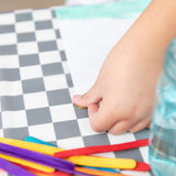 A hand places a coin on a Bumkins Splat Mat: Charcoal Check, with scattered colored craft sticks nearby.