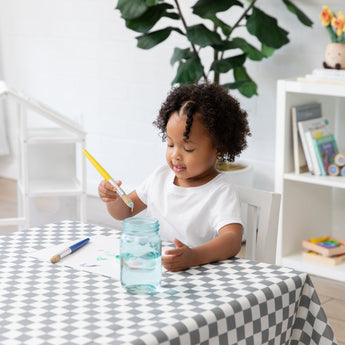 A curly-haired child paints at a table covered with a Bumkins Splat Mat: Charcoal Check, with a plant and books adding charm in the background.