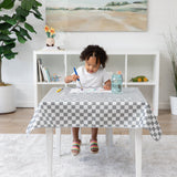 A child paints with watercolors at a table covered in a Bumkins Splat Mat: Charcoal Check, surrounded by toys and a plant.