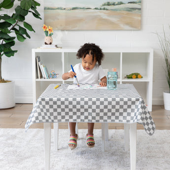 A child paints with watercolors at a table covered in a Bumkins Splat Mat: Charcoal Check, surrounded by toys and a plant.