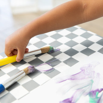 A child paints purple and green streaks on white paper on a Bumkins Splat Mat: Charcoal Check using colorful brushes.