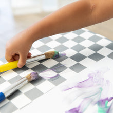 Load image into Gallery viewer, A child paints purple and green streaks on white paper on a Bumkins Splat Mat: Charcoal Check using colorful brushes.
