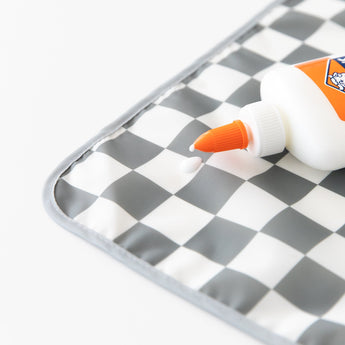Close-up of a white bottle of glue with an orange nozzle dripping onto a Bumkins Splat Mat: Charcoal Check on a white background.