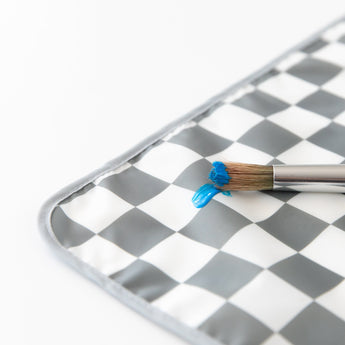 Close-up of a paintbrush with blue paint resting on a Bumkins Splat Mat: Charcoal Check on a white background.