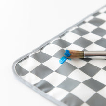 Load image into Gallery viewer, Close-up of a paintbrush with blue paint resting on a Bumkins Splat Mat: Charcoal Check on a white background.
