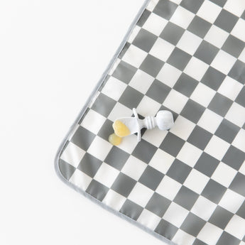 Bumkins Splat Mat: Charcoal Check lays on a white background, with a gray utensil and applesauce on top.