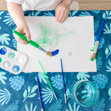 Load image into Gallery viewer, A child paints with green and blue paints on white paper on a Bumkins Splat Mat: Blue Tropic.
