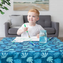Load image into Gallery viewer, A young child paints at a table covered with a Bumkins Splat Mat: Blue Tropic in a cozy living room.

