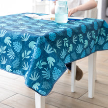 Load image into Gallery viewer, A child paints at a table covered by a Bumkins Splat Mat: Blue Tropic with a blue jar of water.
