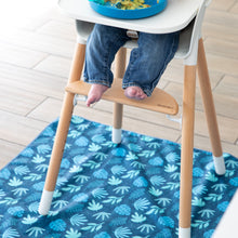 Load image into Gallery viewer, A baby in jeans sits barefoot in a high chair atop a Bumkins Splat Mat: Blue Tropic on a wood floor.
