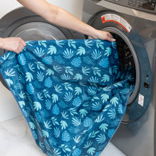 Load image into Gallery viewer, A person places Bumkins Splat Mat: Blue Tropic into an open front-loading washing machine.
