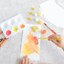 Load image into Gallery viewer, Close-up of a child painting paper on a Bumkins Splat Mat: Wander, surrounded by brushes, a palette, and pom-poms.
