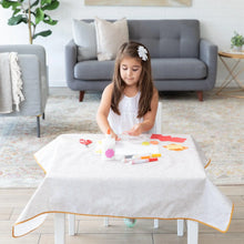 Load image into Gallery viewer, A young girl with a white flower hair clip crafts on a Bumkins Splat Mat: Wander, surrounded by art supplies in a cozy living room.
