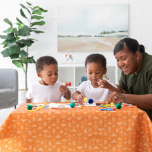 Load image into Gallery viewer, In a sunny room, a woman and two kids enjoy crafts on a bright orange Bumkins Splat Mat: Grounded next to a large plant.
