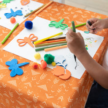 Load image into Gallery viewer, A child draws with markers on a Bumkins Splat Mat: Grounded, surrounded by colorful crafts and pom-poms.
