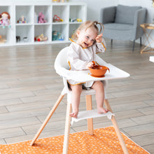 Load image into Gallery viewer, A toddler smiles in a high chair with an orange bowl and spoon, with a Bumkins Splat Mat: Grounded on the floor underneath.
