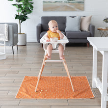 Load image into Gallery viewer, A baby in an yellow bib enjoys a meal in a high chair, the wooden floor protected by the Bumkins Splat Mat: Grounded underneath.
