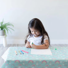 Load image into Gallery viewer, A young girl with long hair sits at a table covered by a Bumkins Splat Mat: Rainbows, coloring on paper with markers.
