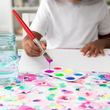 Load image into Gallery viewer, A child paints with watercolors using a brush, while a Bumkins Splat Mat: Watercolor protects the table.

