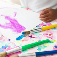 Load image into Gallery viewer, A child paints with bright colors, surrounded by brushes, on a stain-resistant Bumkins Splat Mat: Watercolor.
