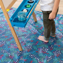 Load image into Gallery viewer, A child stands barefoot on a Bumkins Splat Mat: Animal Prints next to a painting easel with jars of paint.

