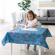 Load image into Gallery viewer, A child paints at a table covered with a Bumkins Splat Mat: Animal Prints; a plant and couch line the background of the bright room.
