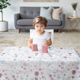 A toddler with short brown hair and a pink bow plays with two small pink sandcastle toys at a table covered by a Bumkins Splat Mat: Floral.