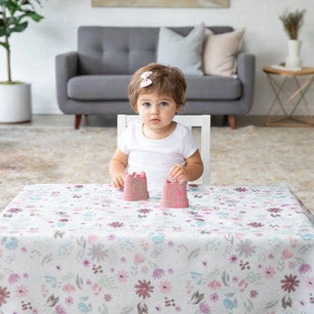 A toddler with short brown hair and a pink bow plays with two small pink sandcastle toys at a table covered by a Bumkins Splat Mat: Floral.