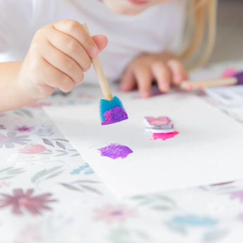 Close-up of a child painting with a blue sponge brush on a Bumkins Splat Mat: Floral.