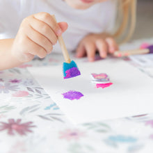 Load image into Gallery viewer, Close-up of a child painting with a blue sponge brush on a Bumkins Splat Mat: Floral.
