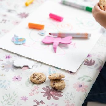 Close-up of a child drawing with markers on a Bumkins Splat Mat: Floral, with cookies nearby.