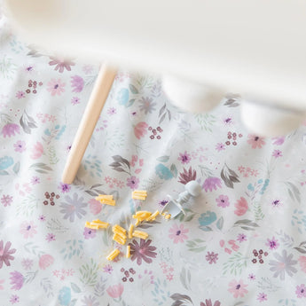 Bumkins Splat Mat: Floral lays under a high chair, covered in macaroni and cheese with a gray silicone utensil nearby.